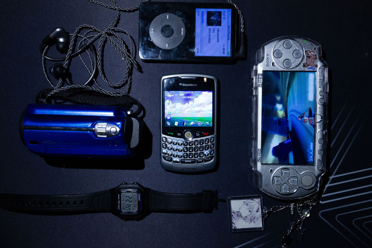 A set of devices laid out on a table. In the center is a BlackBerry Curve, with a PSP, a JVC camcorder, a 5th gen iPod, and a Casio watch around it